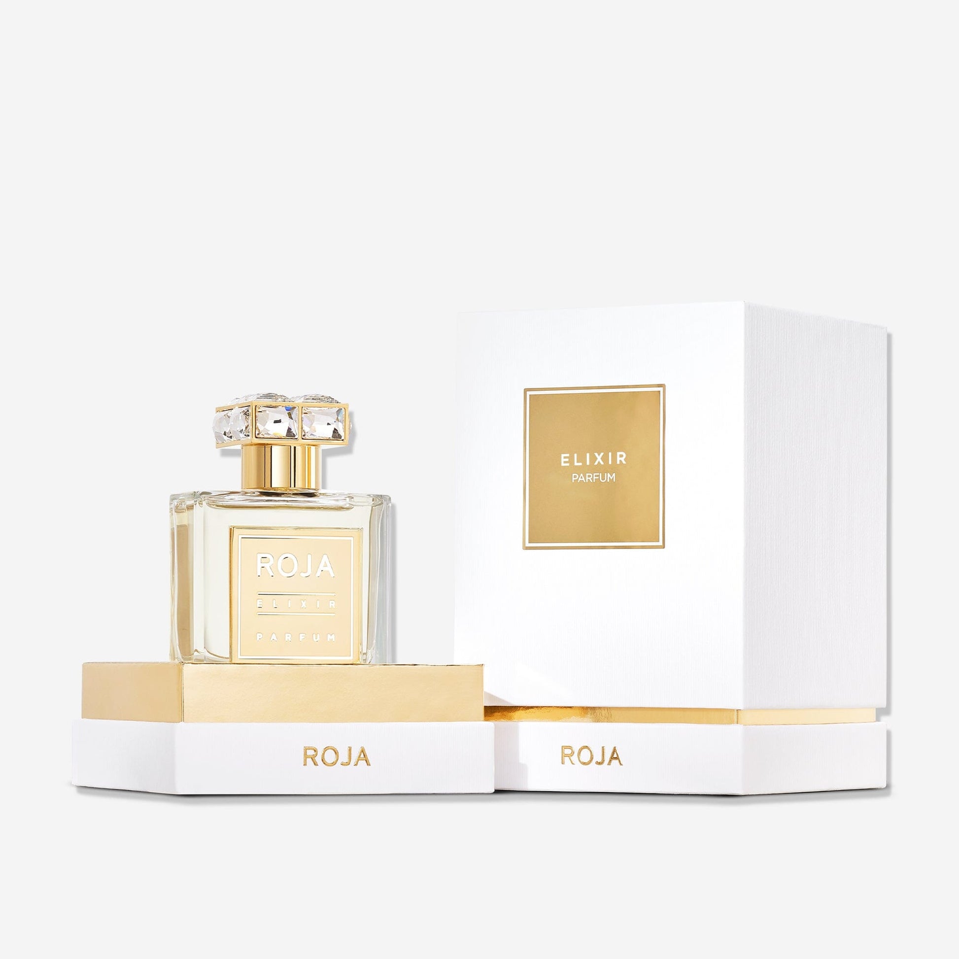 A bottle of Roja Parfums Elixir Pour Femme, featuring a gold label and decorative cap on a white base with "ROJA" in gold, is displayed beside its white and gold box. This ethereal fragrance blends vanilla, amber, and musk notes.