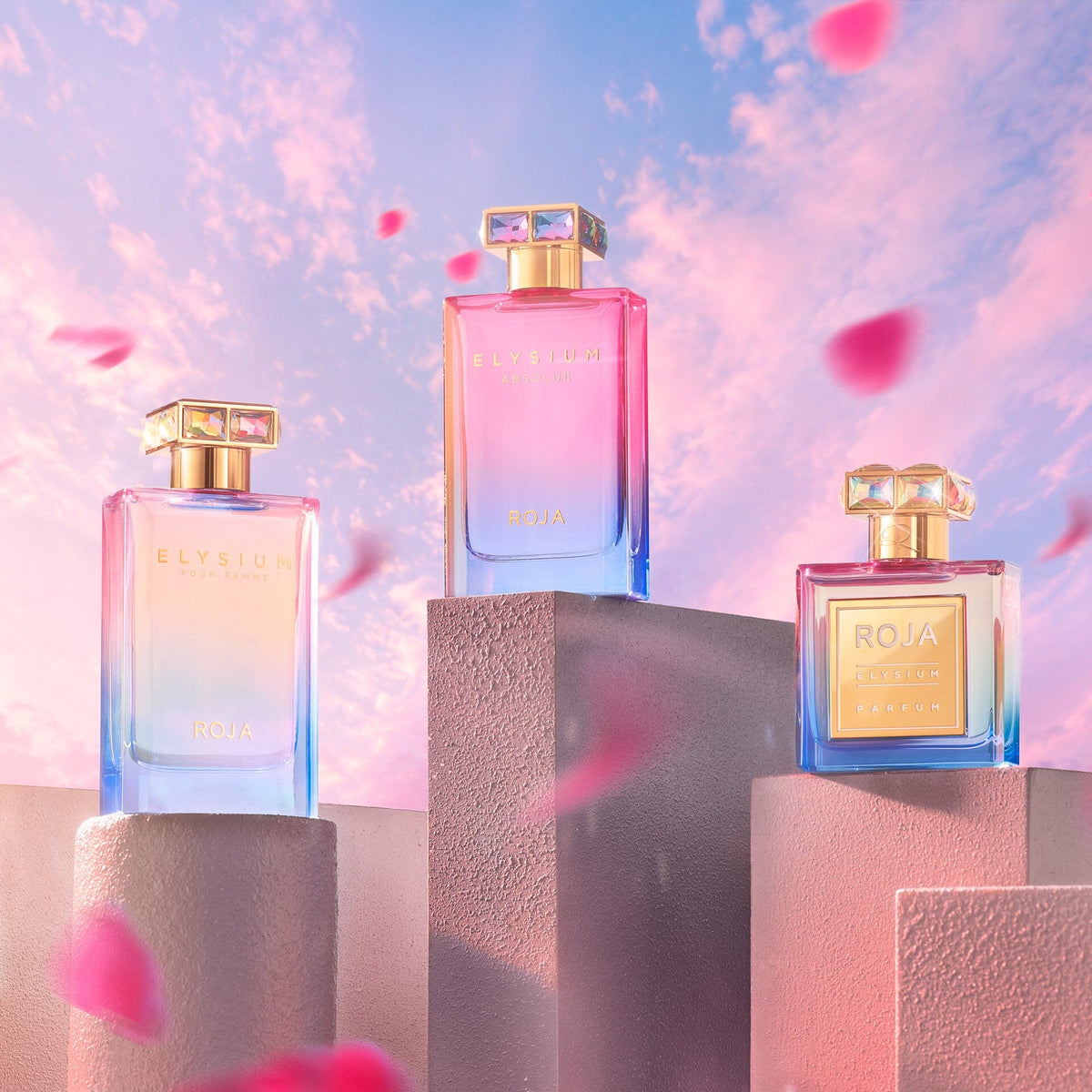 Three vibrant Roja Parfums Elysium Absolue bottles displayed on pastel blocks against a pink and blue sky, with soft pink flower petals drifting in the foreground.