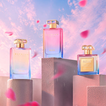 Three vibrant Roja Parfums Elysium Absolue bottles displayed on pastel blocks against a pink and blue sky, with soft pink flower petals drifting in the foreground.