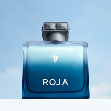 Against a light blue sky, the ROJA Parfums Elysium Eau Intense perfume features a blue glass bottle with a geometric silver cap and sleek white text.