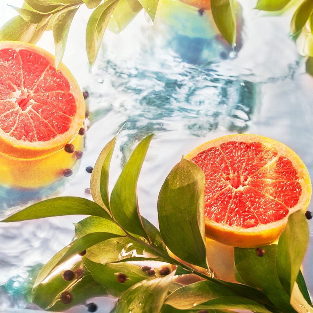 Freshly sliced grapefruits sit among vibrant green leaves and peppercorns on a glossy surface, sunlight casting highlights—an image echoing the invigorating essence of Elysium Eau Intense by Roja Parfums.