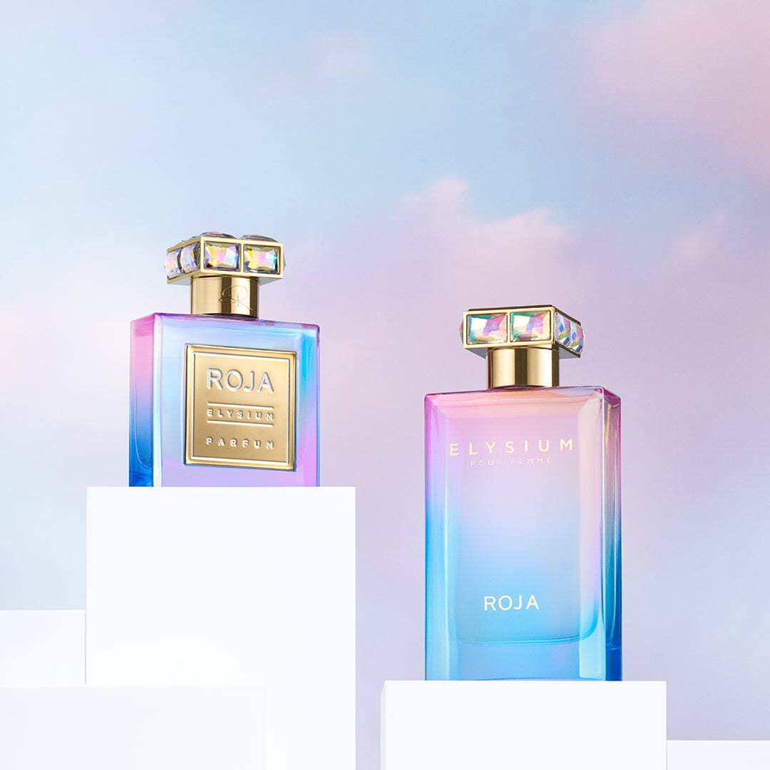 Two elegant Elysium Pour Femme bottles by Roja Parfums sit on white pedestals against a pastel sky, their iridescent blue, pink, and purple hues shimmering. Each features a jeweled facet cap and holds an ethereal blend of scents.