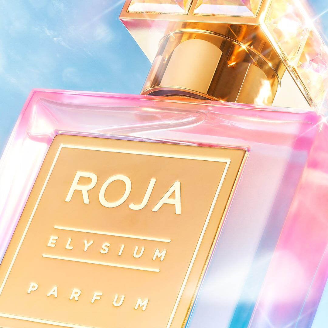 A close-up of an Elysium Pour Femme perfume bottle by Roja Parfums against a blue sky showcases its pink-tinted glass and gold label, resembling an ethereal blend. A chunky, reflective cap crowns it elegantly as sunlight casts a divine goddess glow around the edges.