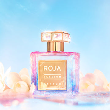 The perfume bottle, labeled Elysium Pour Femme by Roja Parfums, with its jeweled cap, sits among soft white flowers under a bright blue sky. This ethereal blend creates a dreamy and luxurious atmosphere, ideal for invoking the essence of a divine goddess.
