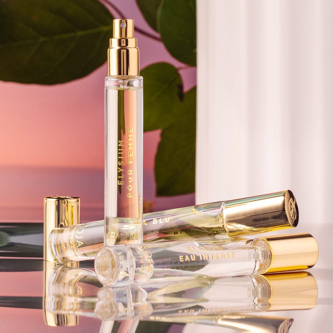 Three elegant Elysium Pour Femme bottles by Roja Parfums, with gold caps and labels, are arranged on a reflective surface among green leaves and a soft pink gradient—two lying down and one upright—exuding an ethereal allure.