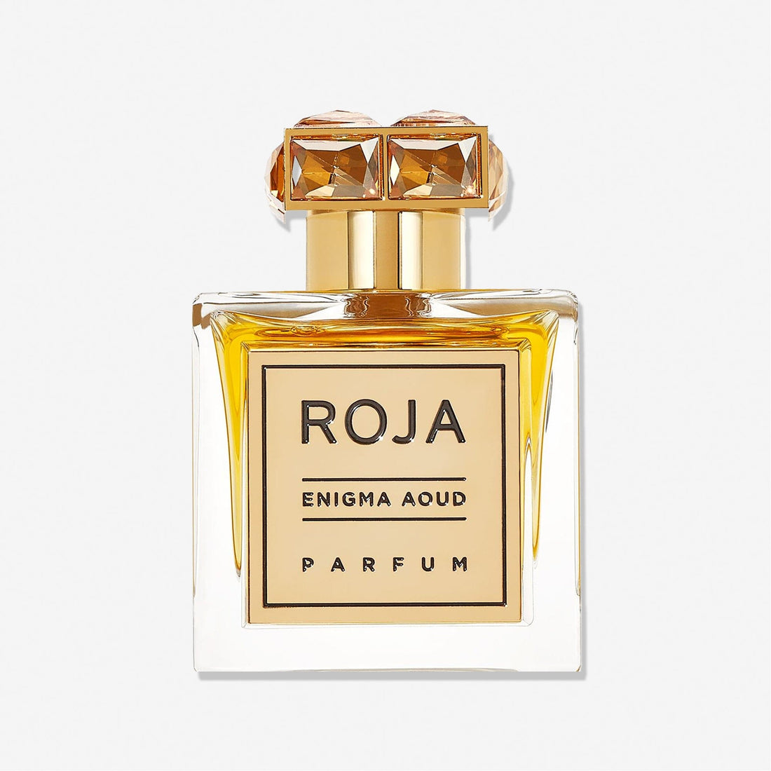 A clear glass perfume bottle of Enigma Aoud by Roja Parfums, featuring golden accents and a crystal-like gold cap, filled with amber-hued scent, is displayed against a plain white background.