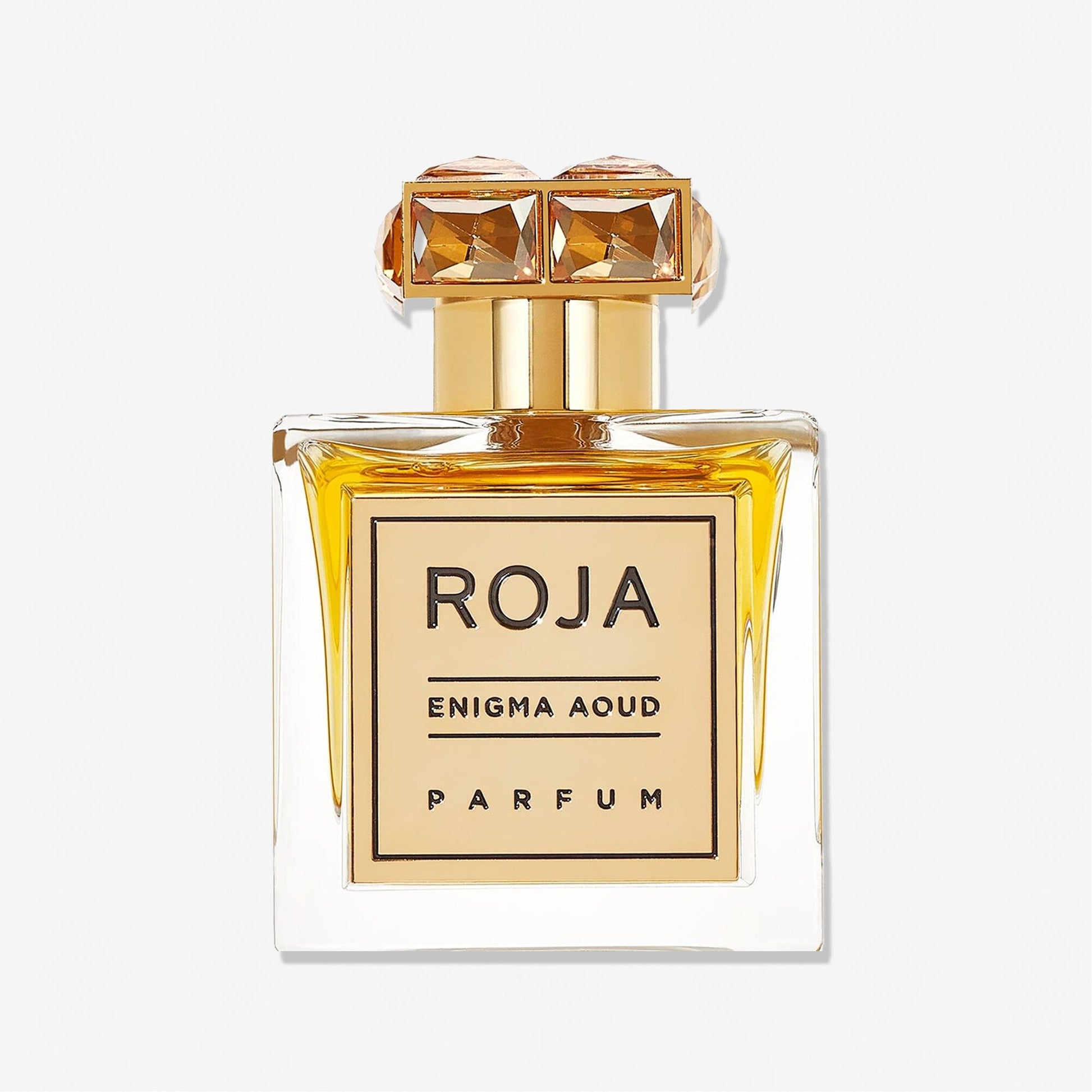 A clear glass perfume bottle of Enigma Aoud by Roja Parfums, featuring golden accents and a crystal-like gold cap, filled with amber-hued scent, is displayed against a plain white background.