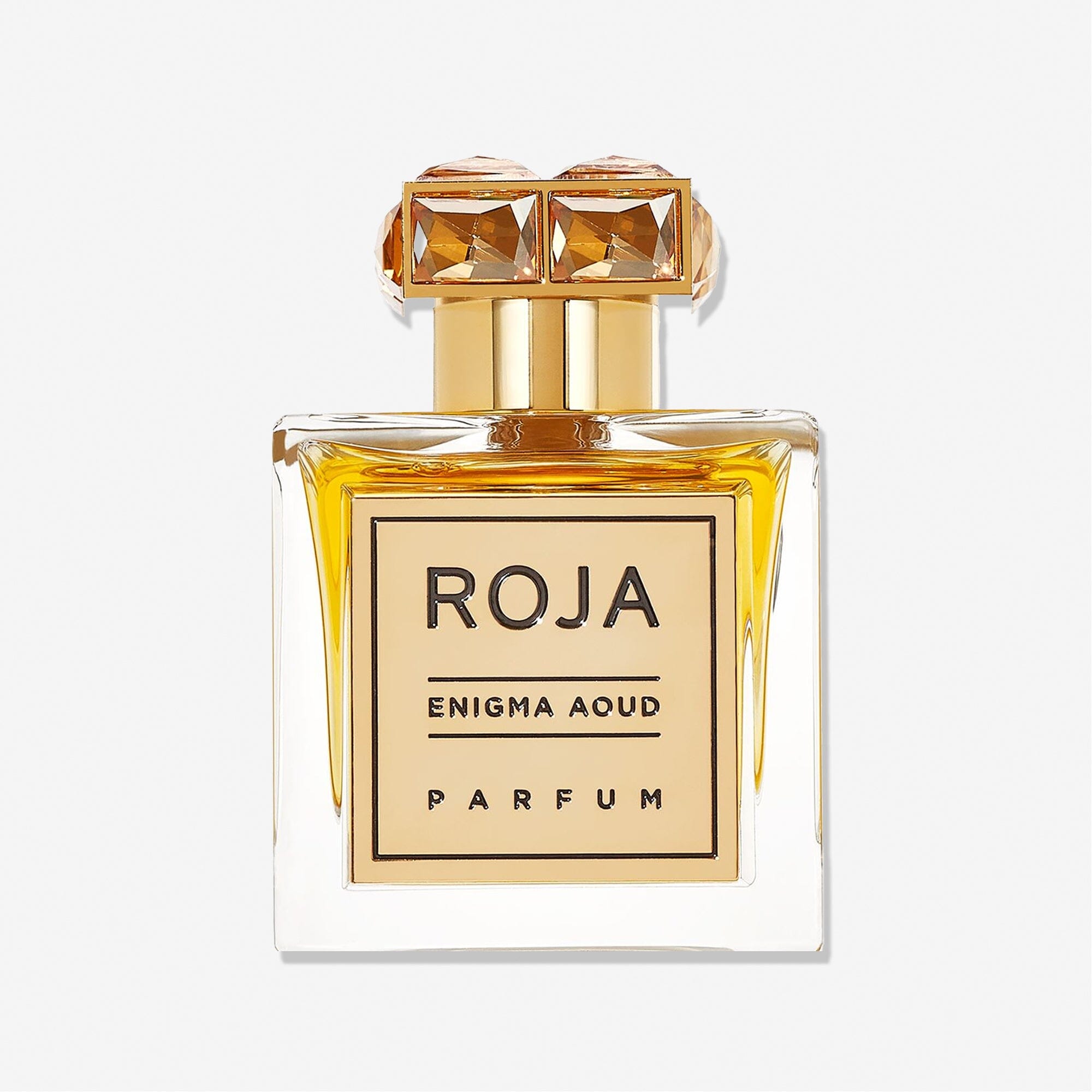 A clear glass perfume bottle of Enigma Aoud by Roja Parfums, featuring golden accents and a crystal-like gold cap, filled with amber-hued scent, is displayed against a plain white background.