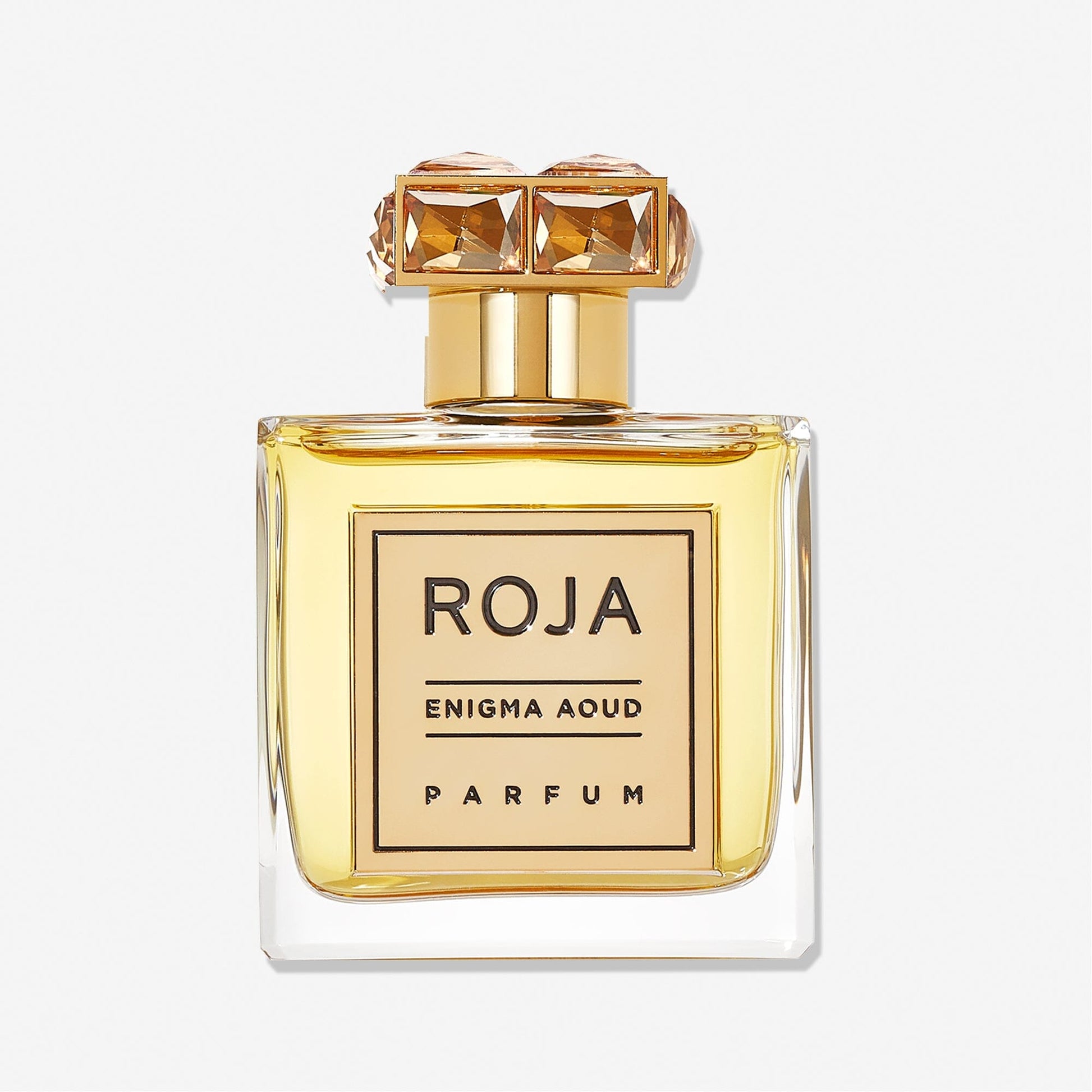 A clear glass bottle filled with golden liquid, labeled Enigma Aoud by Roja Parfums, features a gold cap topped with two jeweled squares—a stunning presentation of this luxurious Arabian fragrance.