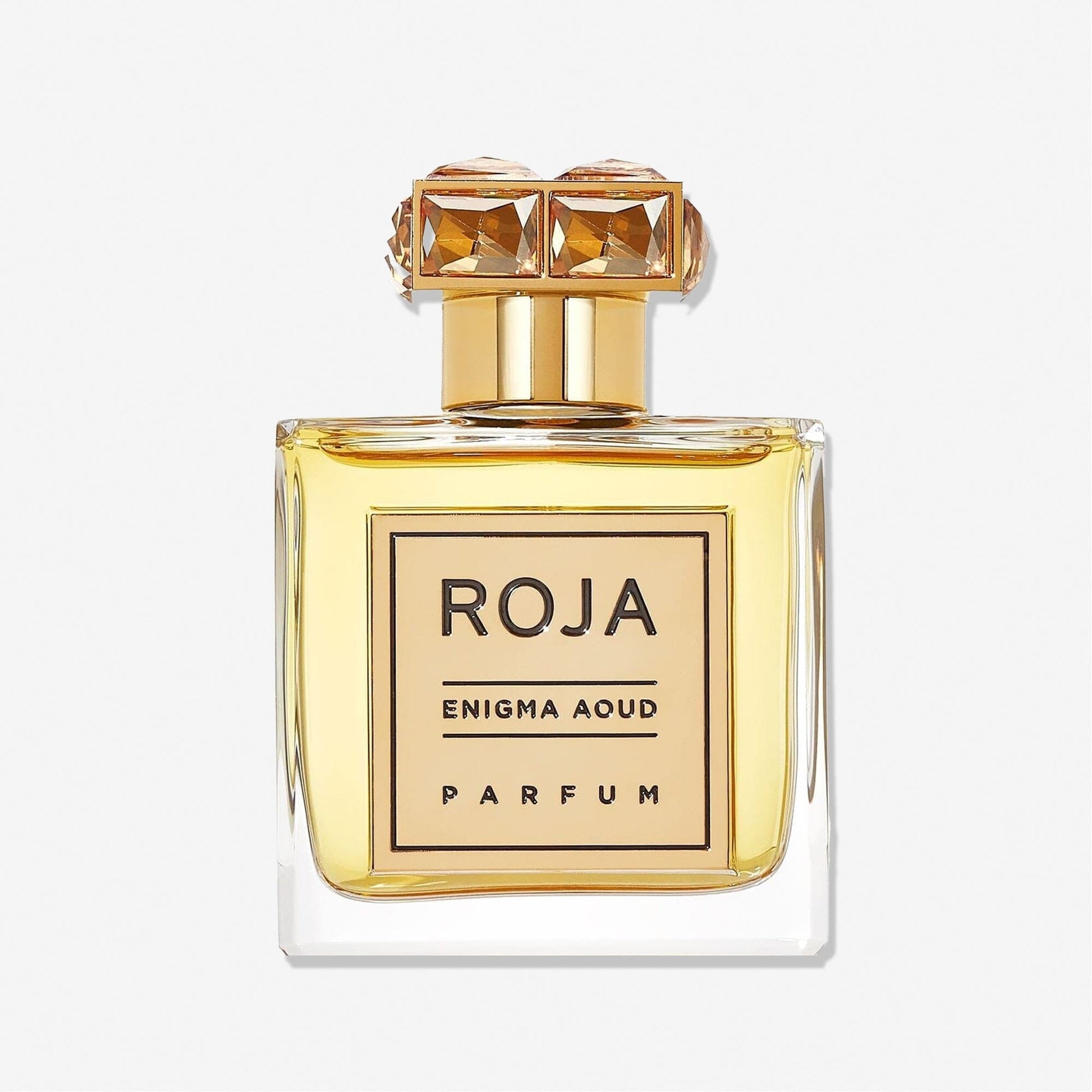 A clear glass bottle of Enigma Aoud by Roja Parfums, featuring a gold cap with two square gem-like accents and a gold label with black text. The radiant golden yellow liquid inside highlights this exquisite fragrance.