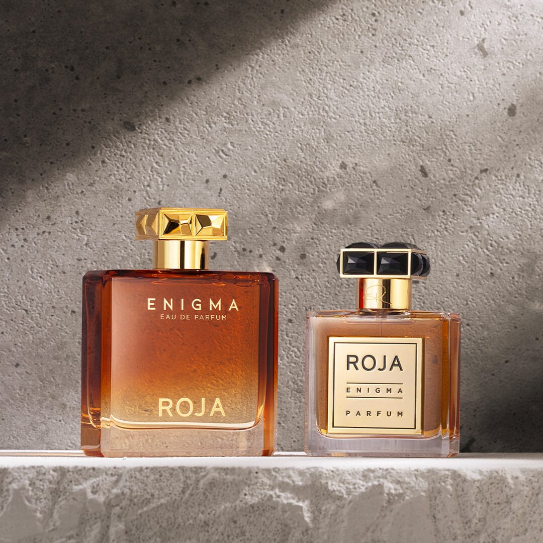 Two square perfume bottles from the Roja Parfums Enigma Gift Set are displayed on a textured gray surface. Both feature gold accents and liquid inside—one larger with a gold cap, the other smaller with a black and gold cap.