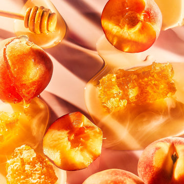 Enigma Pour Femme by Roja Parfums features an intricate blend of fresh peaches, golden honeycomb, and citrus zest. A honey dipper rests among these elements, all bathed in warm natural light on a soft pink surface, creating rich shadows.