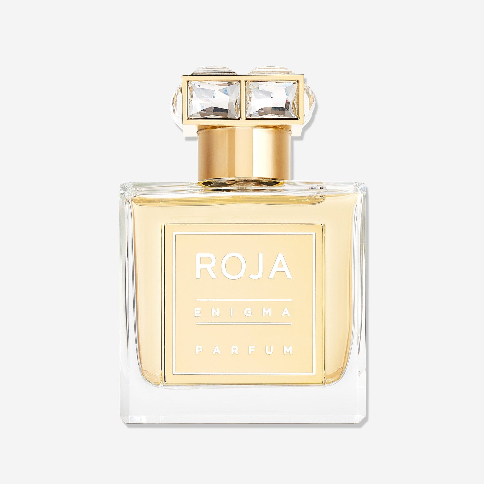 A clear glass bottle of Enigma Pour Femme by Roja Parfums, featuring an ambergris scent, with a gold label and a decorative gold cap set with three square crystal gems, displayed against a plain white background.