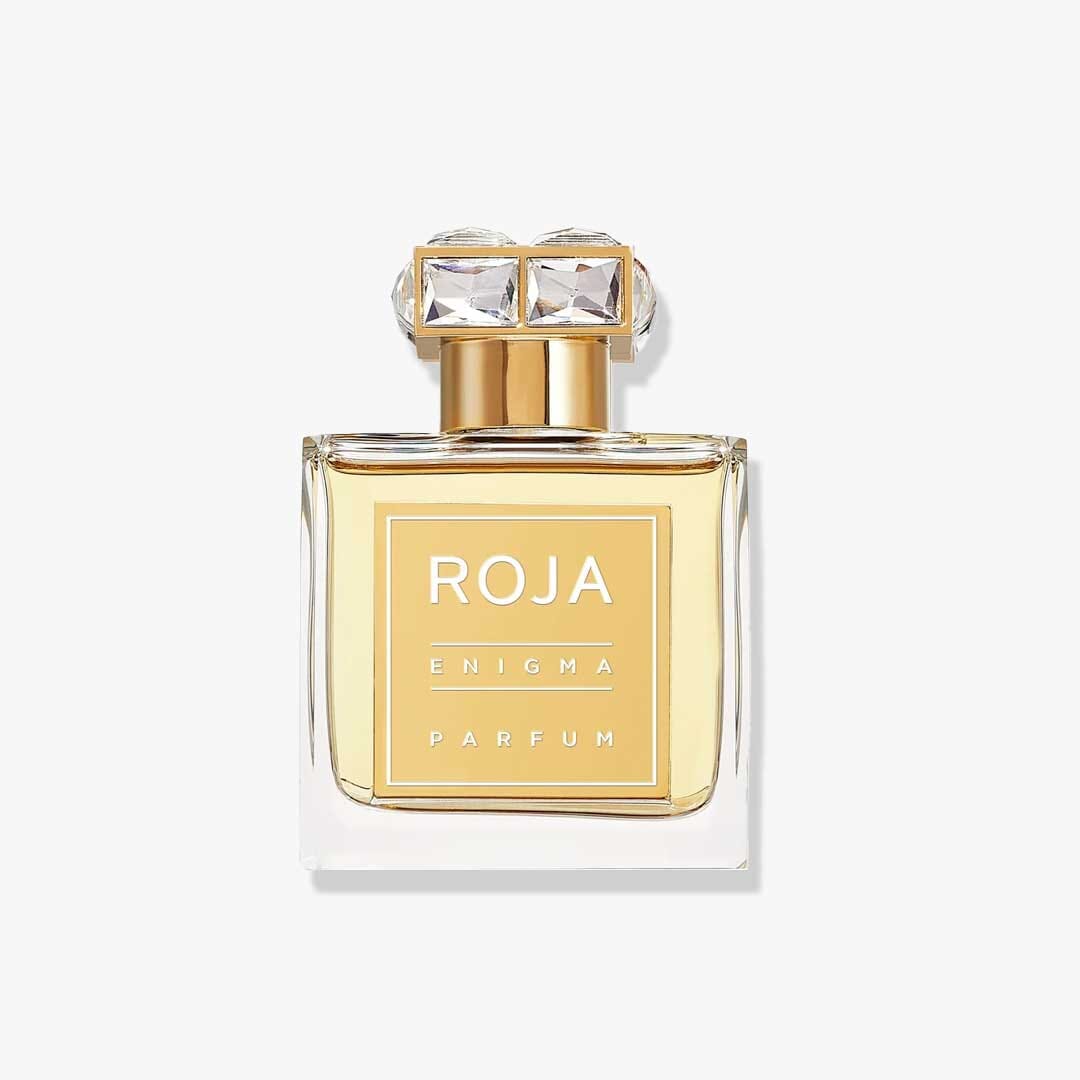 A clear glass bottle of Roja Parfums Enigma Pour Femme, featuring a gold label and cap with two crystal-like accents, suggests notes of citrus and sandalwood, displayed on a plain white background.