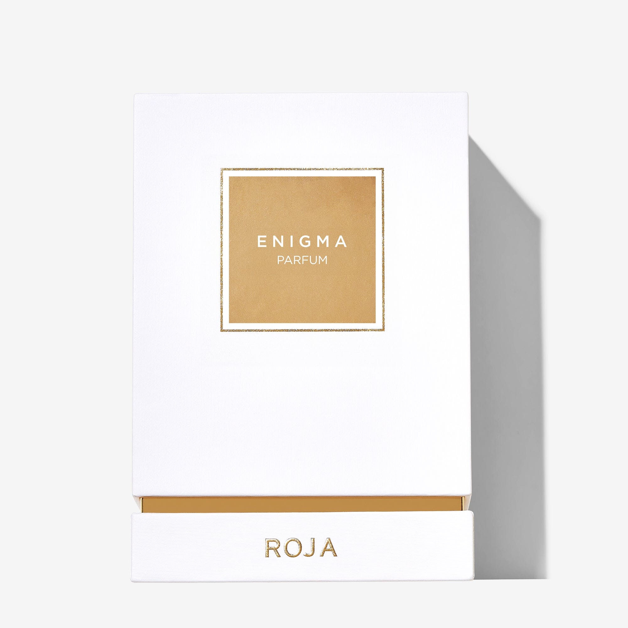 A white rectangular box with a gold-bordered label reading ENIGMA PARFUM and ROJA hints at the luxurious scent of Enigma Pour Femme by Roja Parfums, revealing notes of peach, ambergris, and musk inside its gold interior.