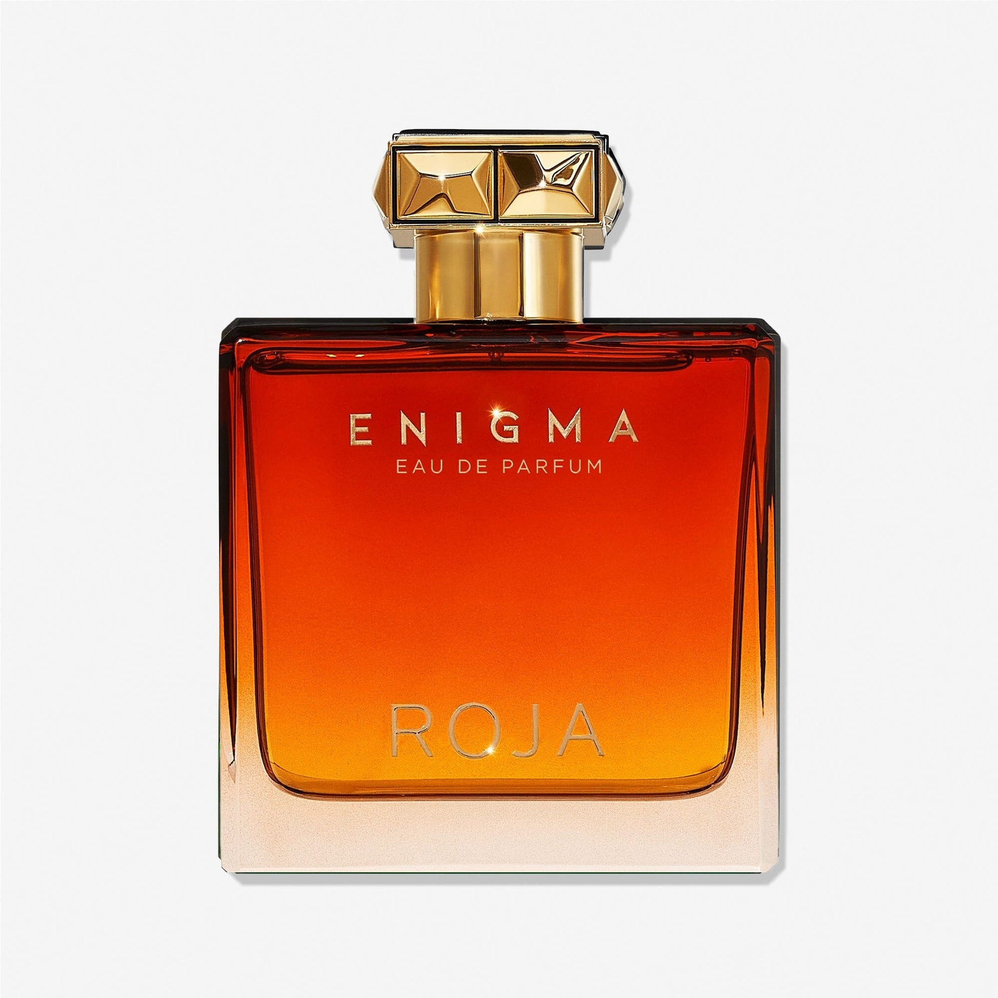 A rectangular glass bottle featuring a gradient amber-to-clear color, gold cap with geometric design, and gold “Enigma Pour Homme Roja Parfums” text on the front, evoking rich tobacco and smooth vanilla notes.