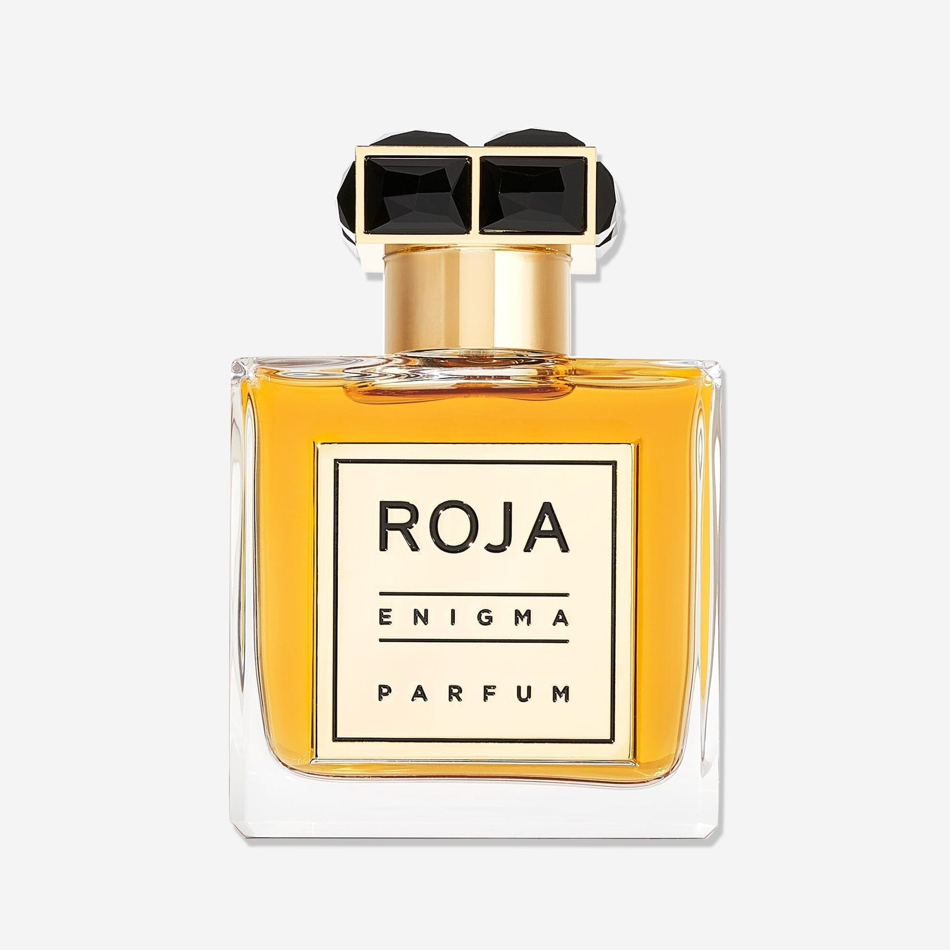 A clear glass bottle of Roja Parfums Enigma Pour Homme holds golden-yellow liquid. The gold cap with two black gem accents complements the label reading ROJA PARFUMS ENIGMA POUR HOMME, evoking the essence of a Cognac-inspired fragrance.