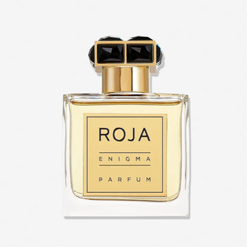 A clear glass bottle of Enigma Pour Homme by Roja Parfums, a men’s fragrance with Tobacco and Cognac notes, displays a gold label and black-and-gold cap. The pale yellow liquid stands out against a plain white background.