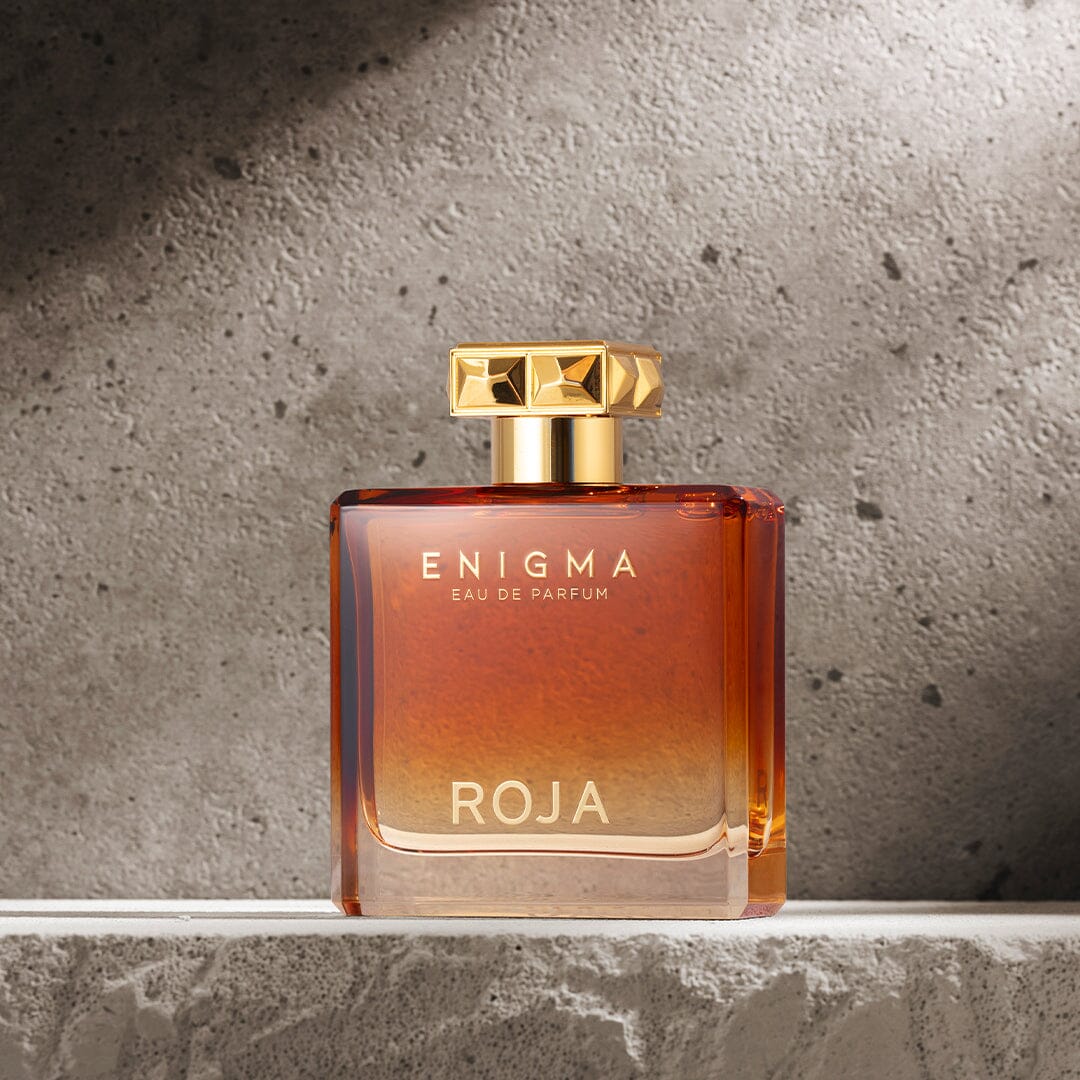 A square glass bottle of Enigma Pour Homme by Roja Parfums, filled with amber-hued liquid, stands on a textured gray stone. The gold cap and lettering evoke the Cognac fragrance and tobacco notes of this luxurious parfum.