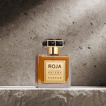 A bottle of Roja Parfums Enigma Pour Homme, featuring amber liquid, a gold label, and a black-gold cap, rests on textured stone, evoking a masculine scent inspired by tobacco and cognac against a gray concrete backdrop.