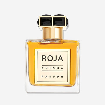 A clear glass bottle of amber-hued Enigma Pour Homme by Roja Parfums features a gold cap with a black, geometric top. The cream label reads ROJA PARFUMS ENIGMA POUR HOMME, holding a rich cognac and tobacco-inspired cologne.