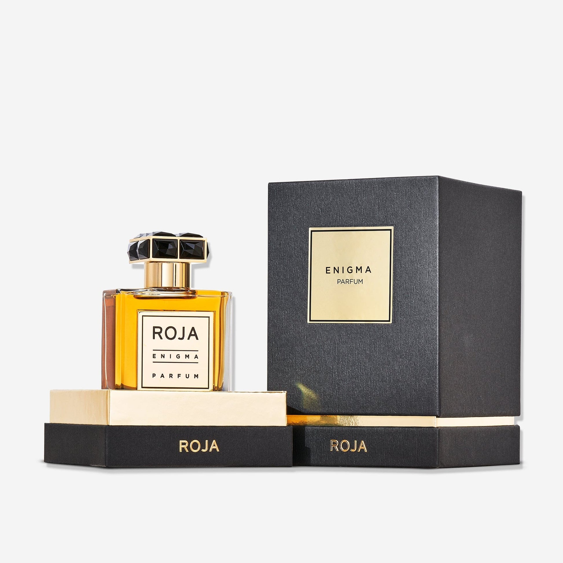 A bottle of Enigma Pour Homme by Roja Parfums, a bold masculine tobacco scent, rests on a gold and black platform next to its matching box—both displaying the product name—against a white background.