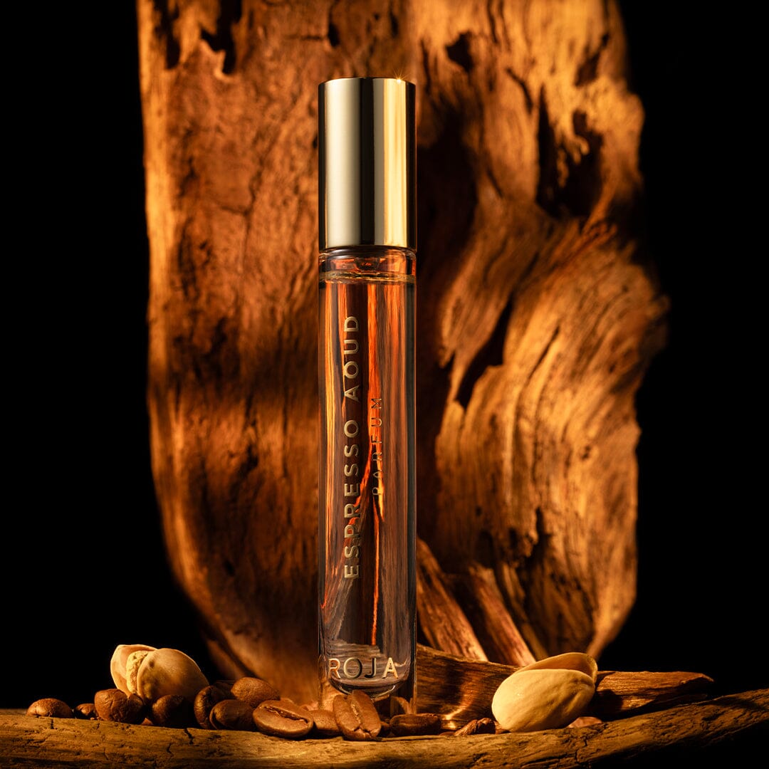 A luxury Roja Parfums Espresso Aoud bottle stands upright on a wooden surface amid coffee beans and pistachios, set against a dramatic wooden backdrop with warm lighting.