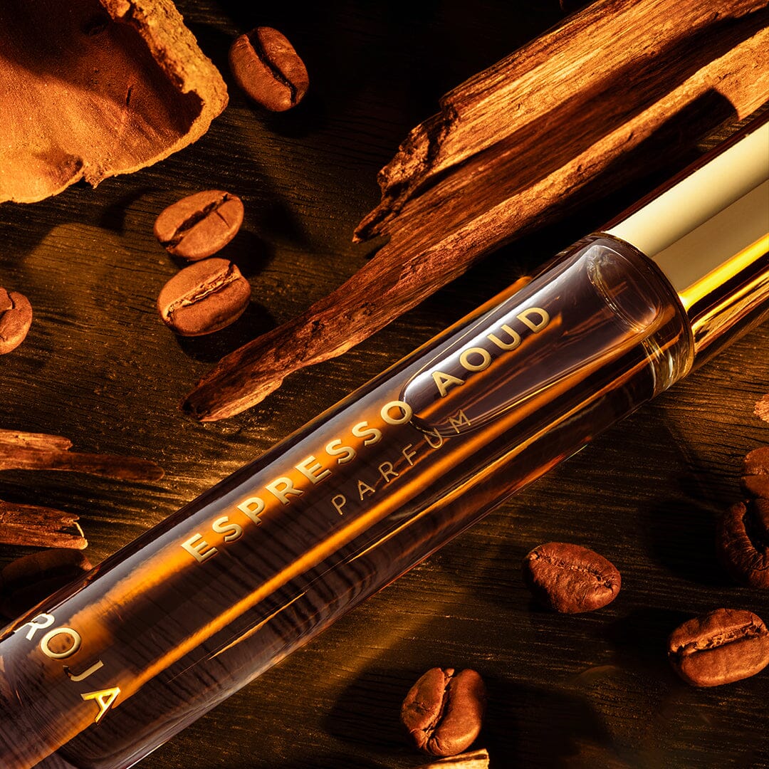 A close-up of a Roja Parfums Espresso Aoud bottle, a luxurious fragrance, rests on wood with coffee beans and wood pieces around it, all illuminated by warm golden light.