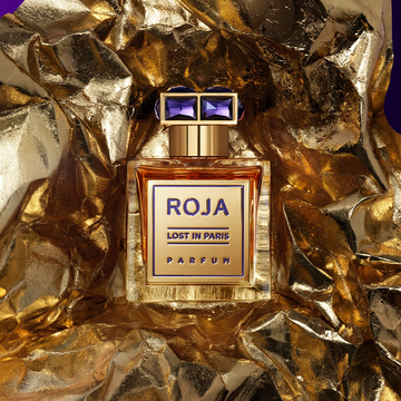 A bottle of Roja Lost in Paris Parfum sits on crumpled gold foil, with a gold cap and purple gem-like accents, creating a luxurious and glamorous display.