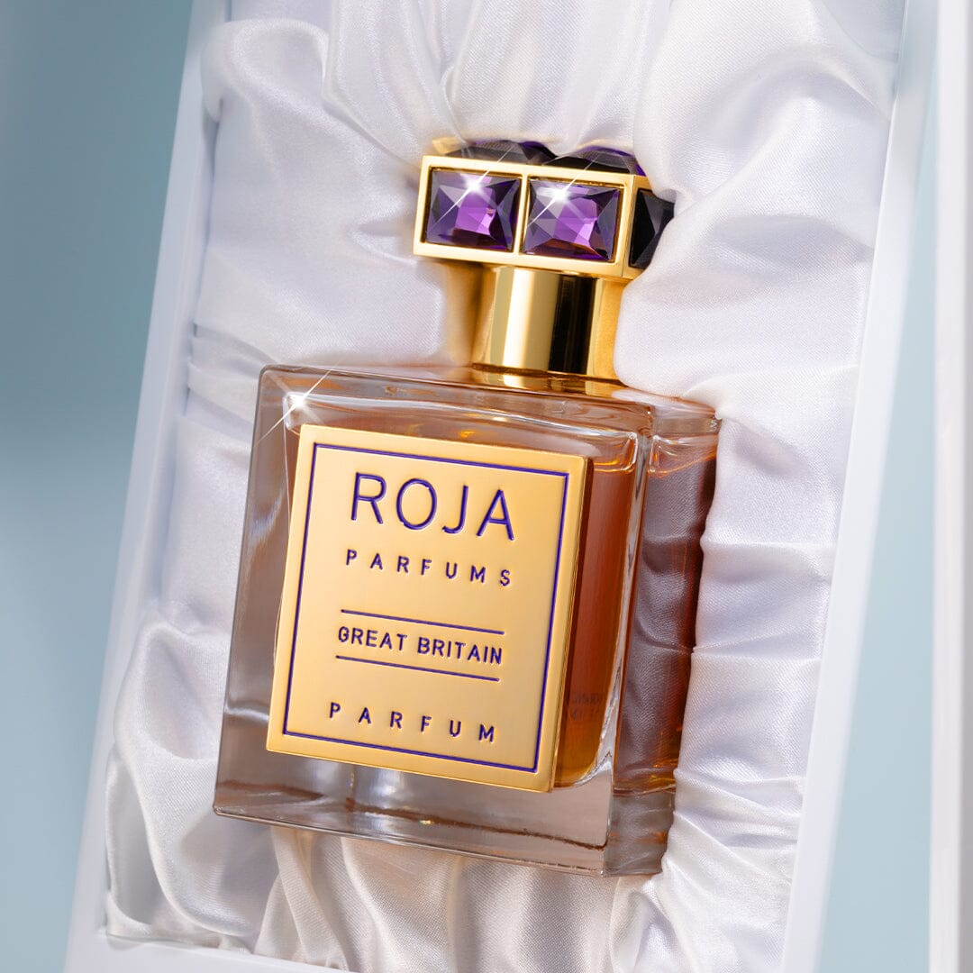 A luxurious bottle of Roja Parfums Great Britain rests on white satin, featuring a gold cap set with purple gems and a gold label with elegant text.