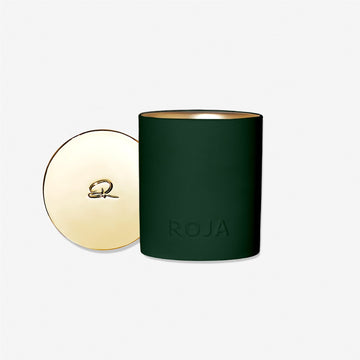 A dark green Harrods Musk Aoud candle by Roja Parfums, with "ROJA" embossed on the front, sits beside a gold-toned lid adorned with a small decorative emblem. The background is plain white.