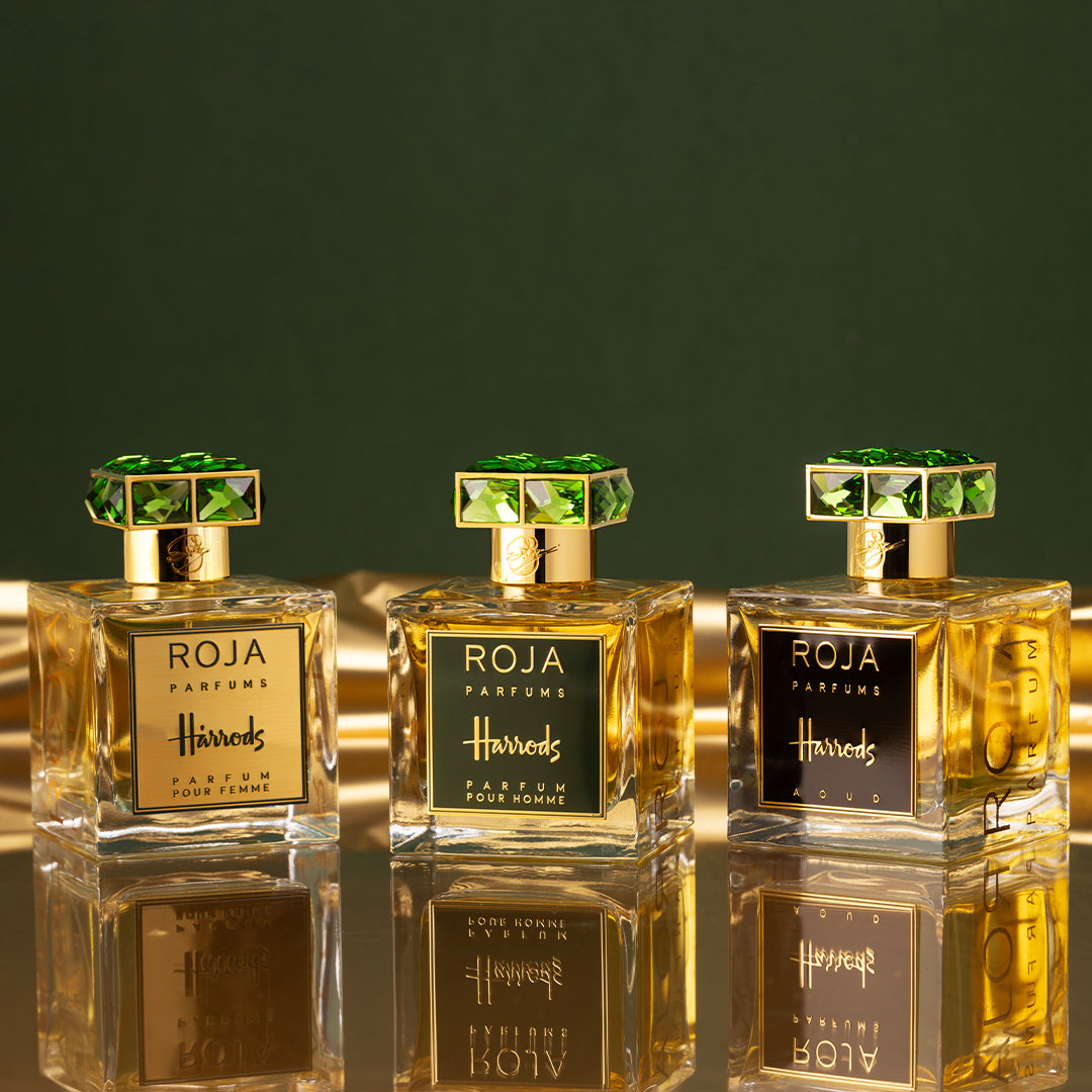Three bottles of Roja Parfums for Harrods are displayed on a reflective gold surface with a green background. Each bottle has a gold label and ornate green gemstone-like cap.