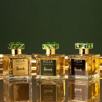Three bottles of Roja Parfums for Harrods are displayed on a reflective gold surface with a green background. Each bottle has a gold label and ornate green gemstone-like cap.