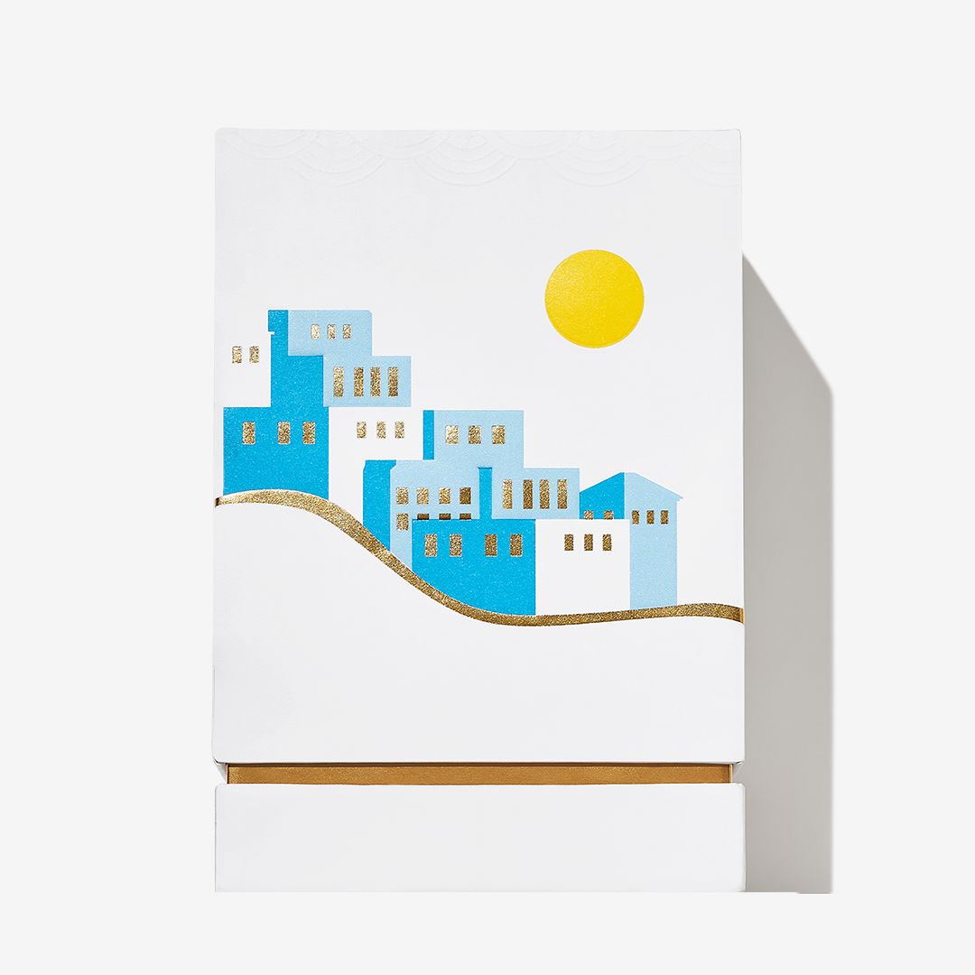 A white box for Roja Parfums Isola Blu features a minimalist design of blue and white hillside buildings, gold window accents, a gold hill line, and a bright yellow sun—evoking citrus freshness on a plain background.