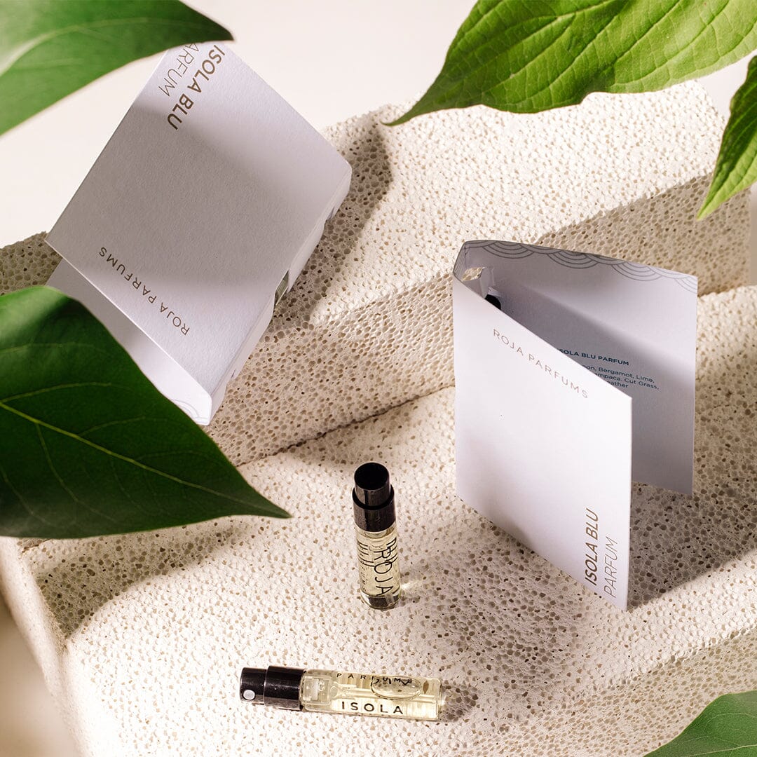 Two Isola Blu Sample (Gift) vials by Roja Parfums rest on a textured white surface, surrounded by green leaves and two open white Roja Parfums booklets in soft natural light.