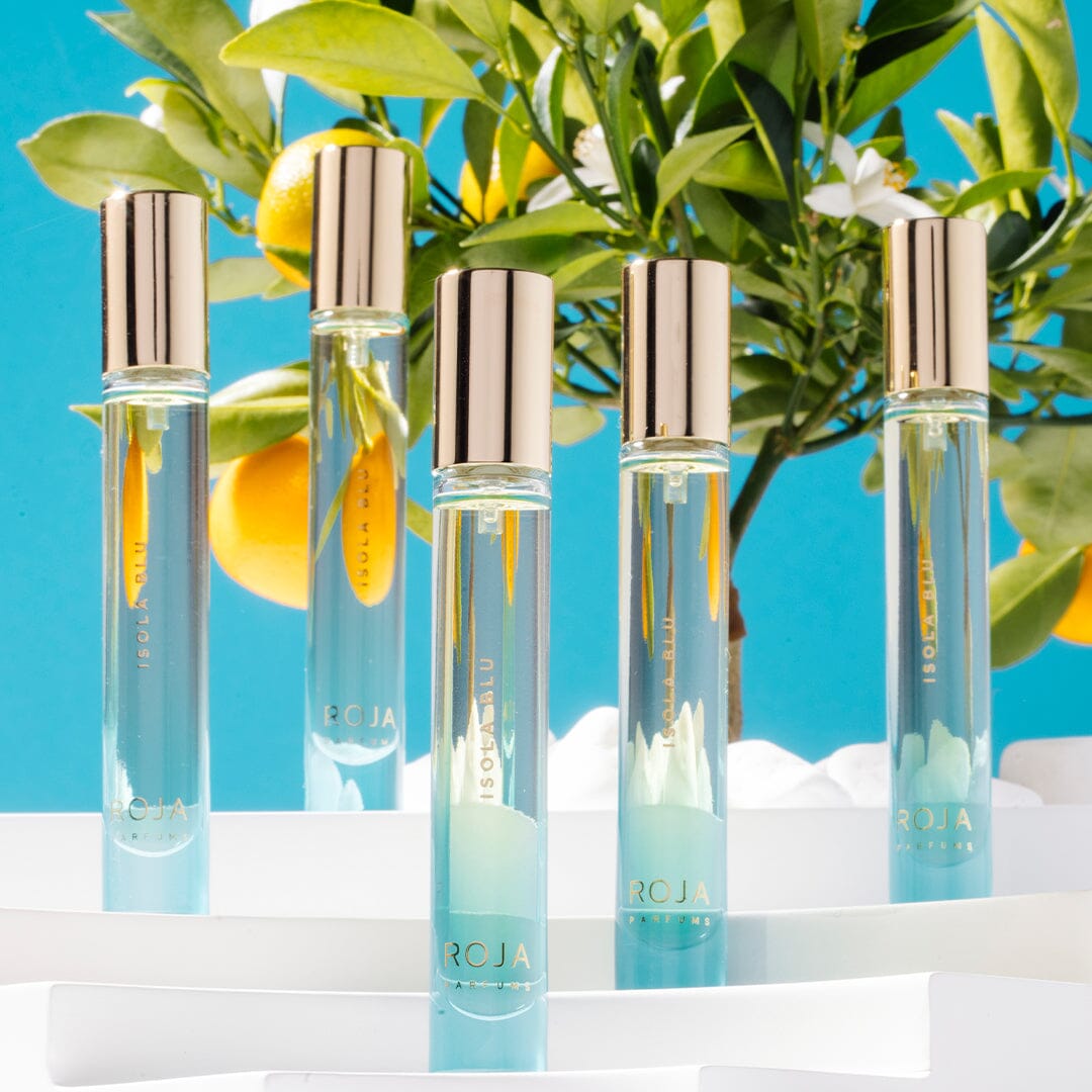 Five elegant Isola Blu perfume bottles by Roja Parfums, topped with gold caps, are displayed on white platforms before a leafy orange tree and blue sky, capturing the luxury and citrus essence of a five-star private villa getaway.
