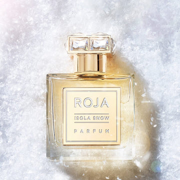 A gold-capped glass bottle of Isola Snow by Roja Parfums rests on a sparkling white, snowy background, with light shimmering off the textured surface.