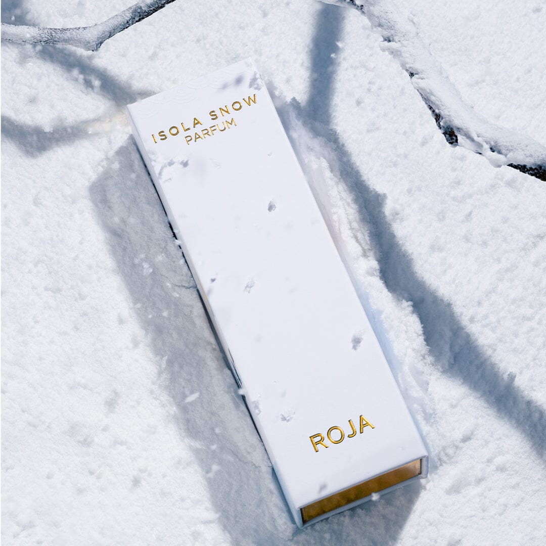 A white rectangular Isola Snow box by Roja Parfums with gold lettering rests on snow beside dark branches, evoking the elegance of a luxury winter escape.