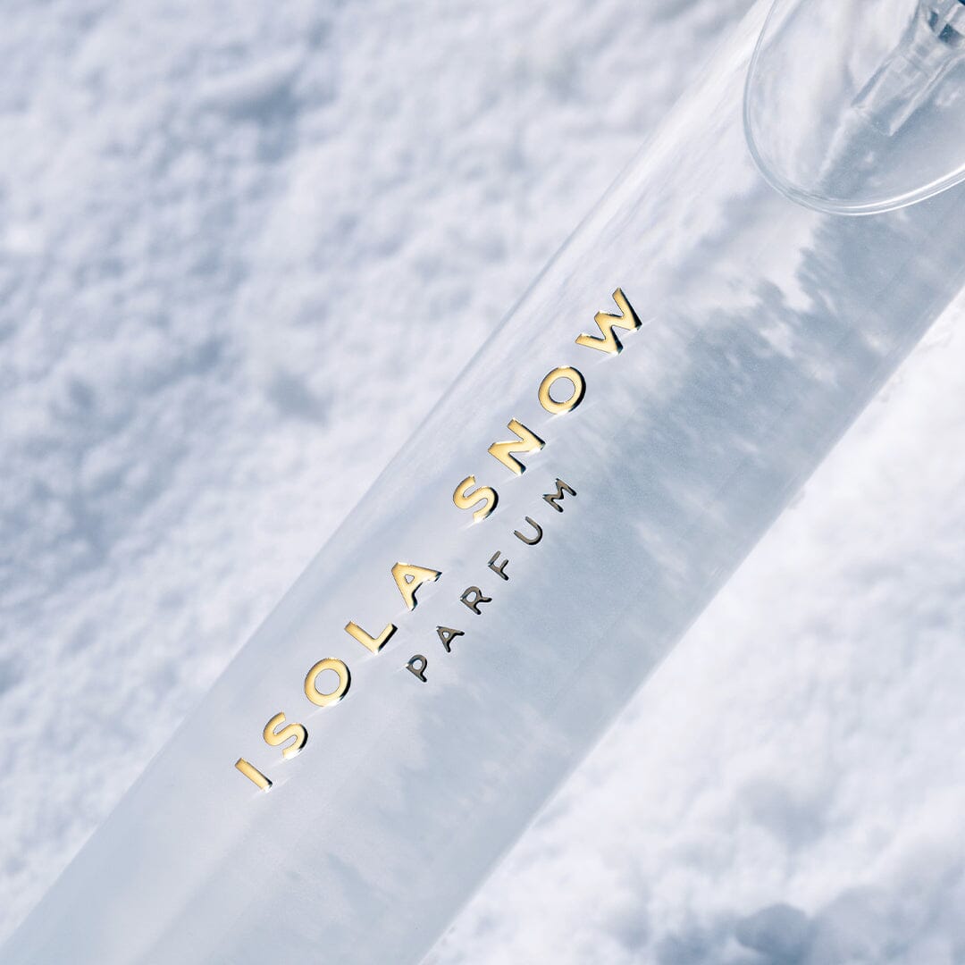 A sleek Isola Snow bottle by Roja Parfums rests on snow, its gold lettering striking against the white background—evoking the serenity of a luxurious island resort spa.