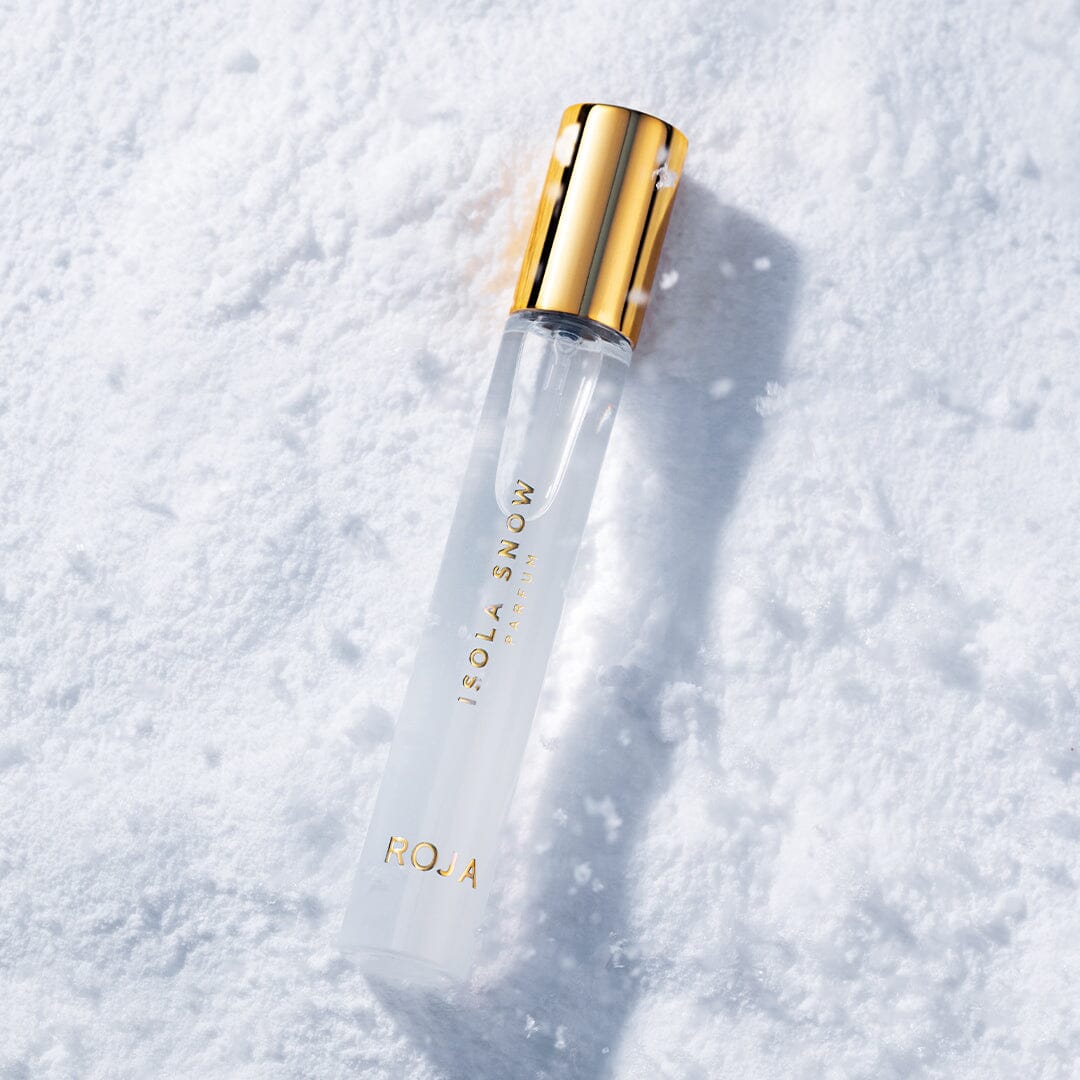 A clear glass perfume bottle with a gold cap labeled ISOLA SNOW by Roja Parfums rests on a snowy white surface, capturing the serene elegance of winter and highlighting the luxurious contrast between glass and snow.