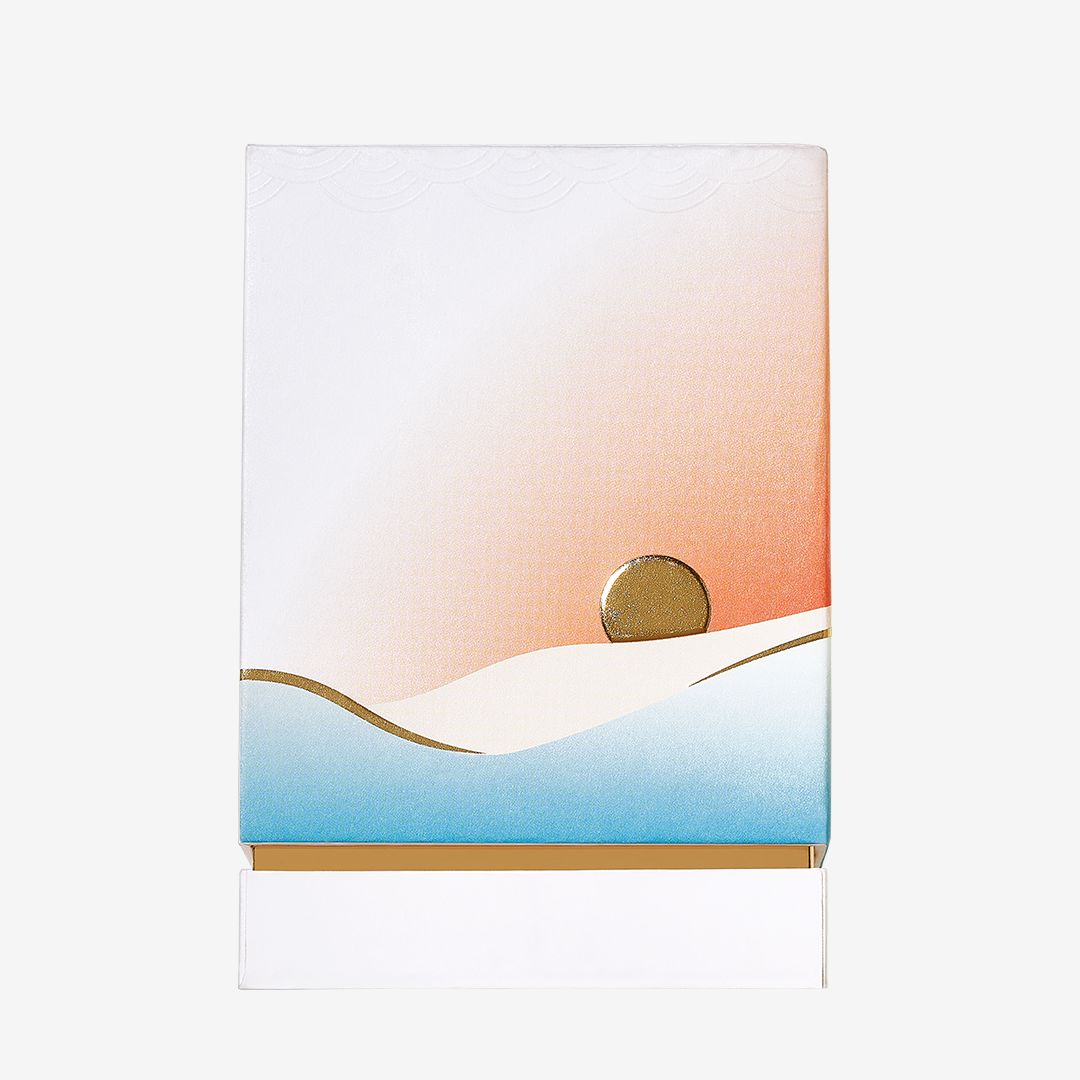 A minimalist illustration on the rectangular Isola Sol packaging by Roja Parfums shows a gold sun over stylized dunes with a gradient sky from white to orange and blue, evoking an island escape. The box is partially open at the bottom.