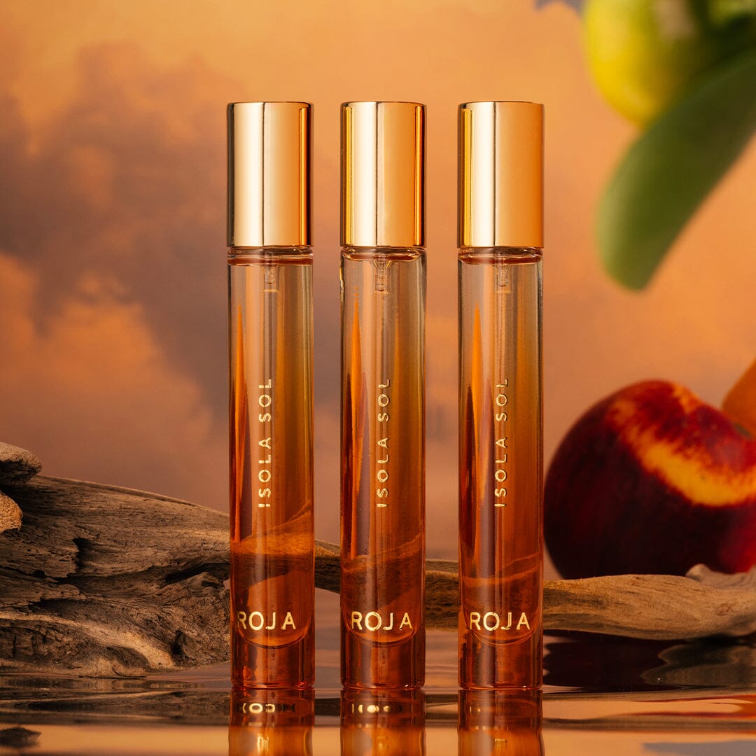 Three elegant perfume bottles labeled ISOLA SOL ROJA stand upright on a reflective surface, with a warm, sunset-colored background featuring a peach and green leaves.