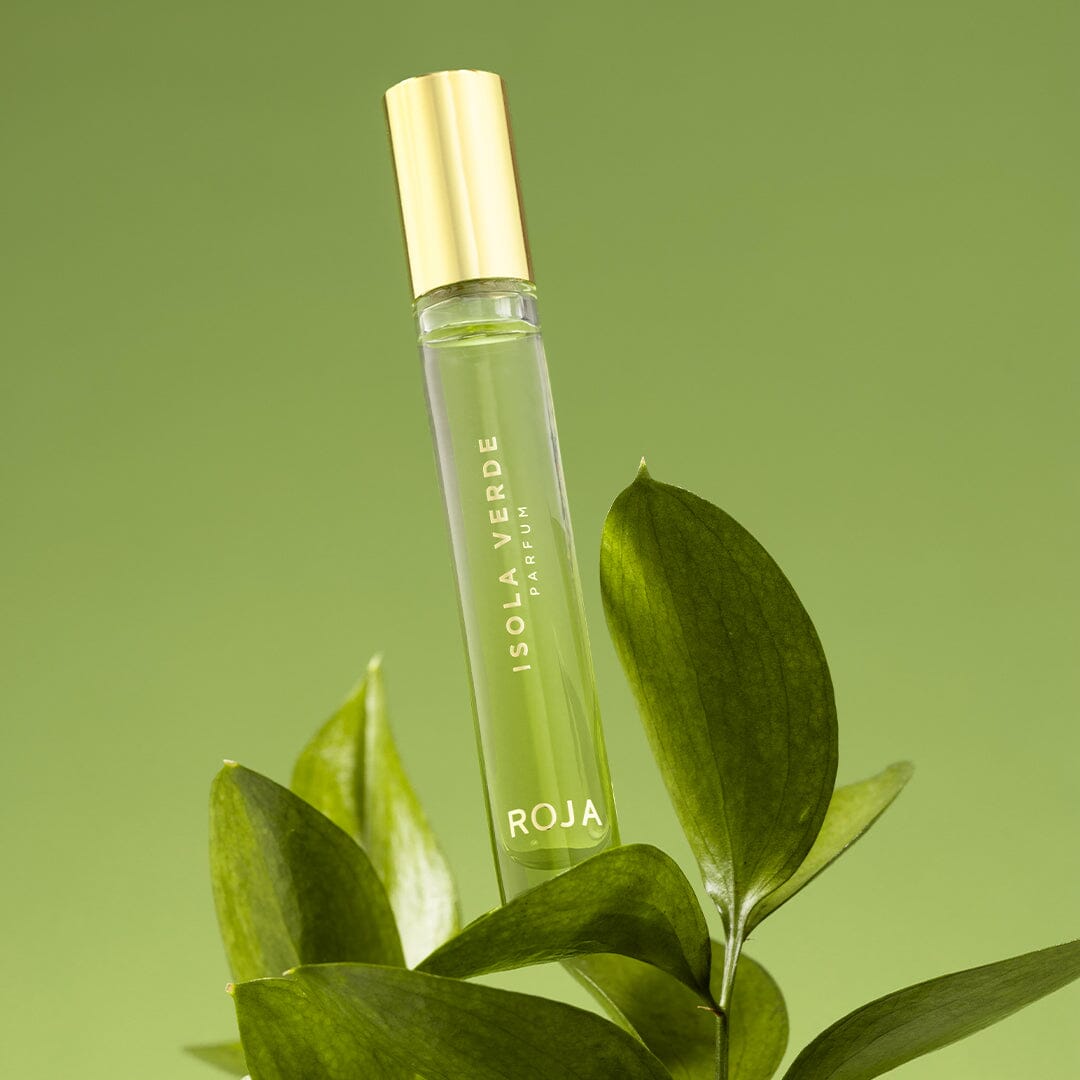 A glass bottle of Roja Parfums Isola Verde, crowned with a gold cap, rests among green leaves, capturing the fragrances exotic floral notes and the allure of turquoise waters against a lush backdrop.