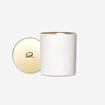 A white cylindrical London candle by Roja Parfums, embossed with ROJA, sits next to its gold-toned lid featuring an R logo—reflecting Londons refined hedonism—all set against a plain white background.