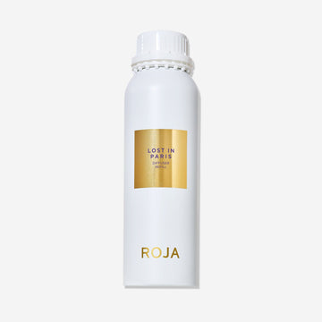 A white cylindrical bottle with a white cap displays a gold label reading Lost in Paris and ROJA London near the bottom. This ROJA London Lost in Paris diffuser captures a gourmand essence. The background is plain white.