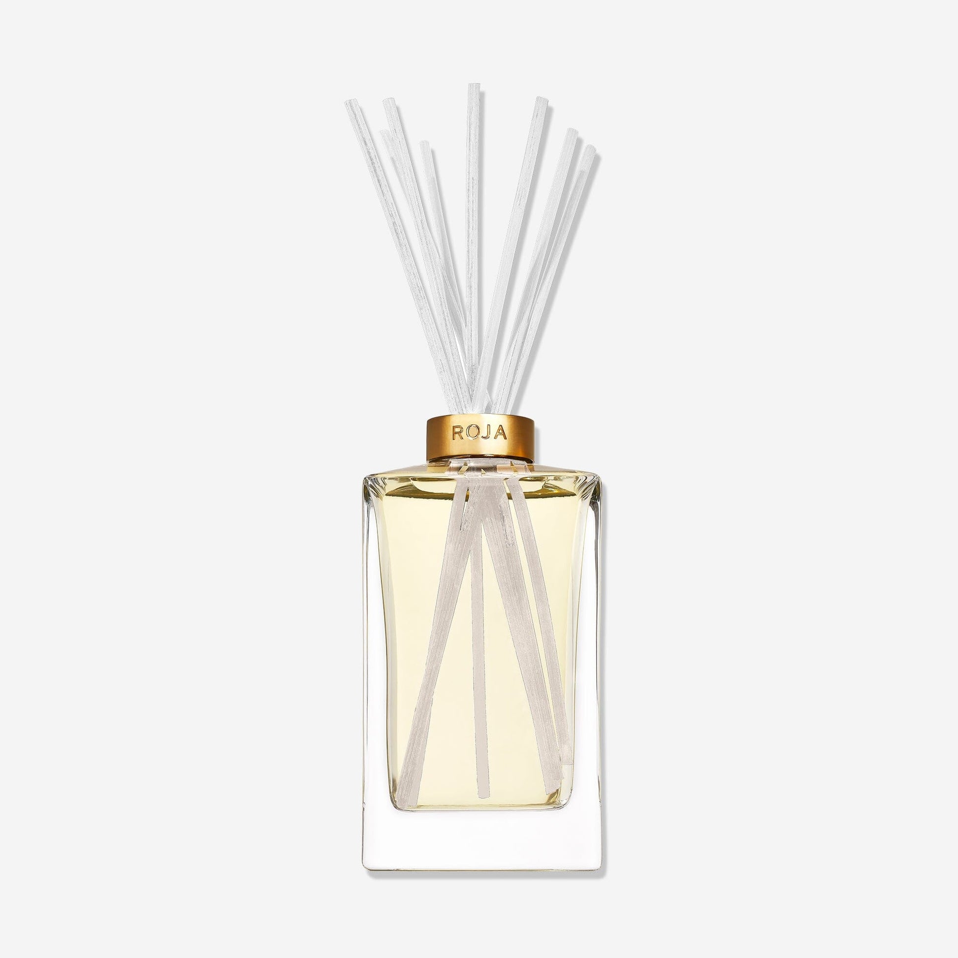 A clear ROJA London Lost in Paris diffuser with light yellow liquid, a gold ROJA collar, and thin white reeds. This gourmand fragrance exudes elegance against a plain white backdrop.