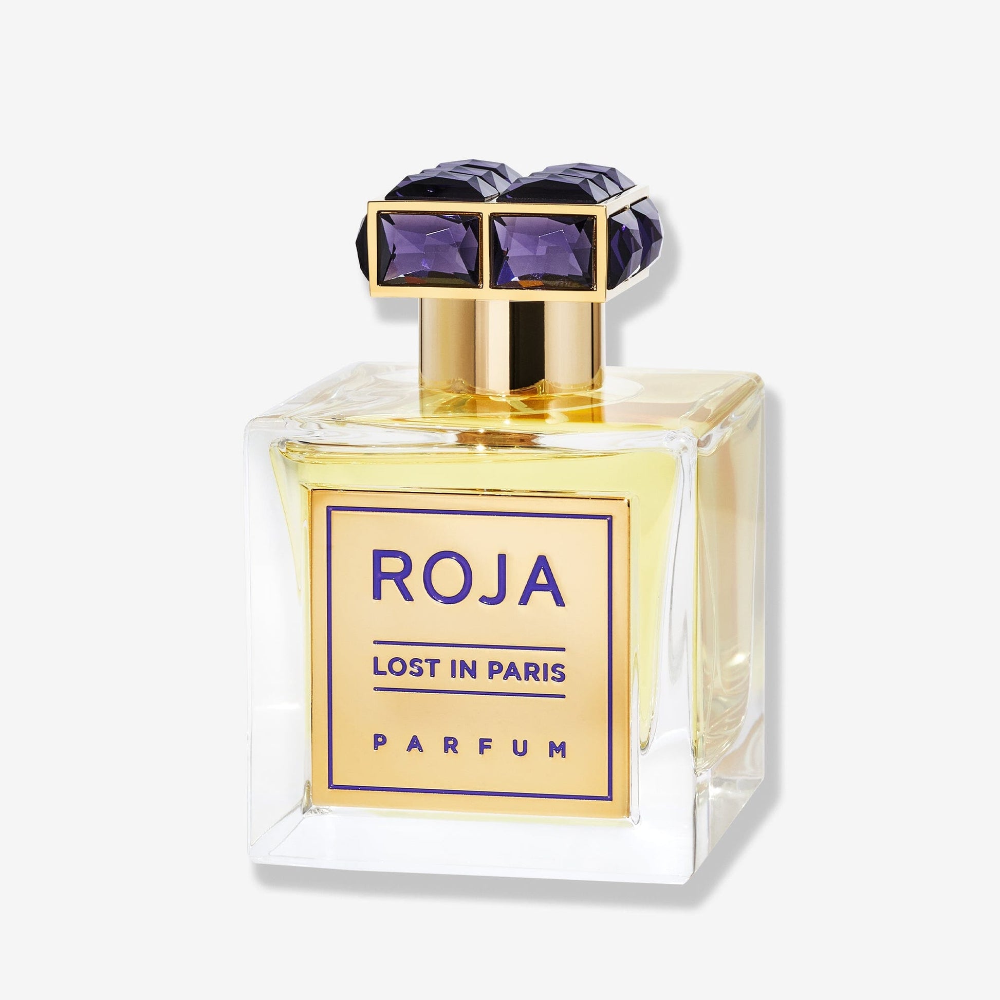 A square glass bottle of Roja Parfums Lost In Paris, a luxury fragrance with golden liquid. It has a gold label and a decorative gold cap set with large, faceted purple gems—capturing the elegance of Parisian high society.