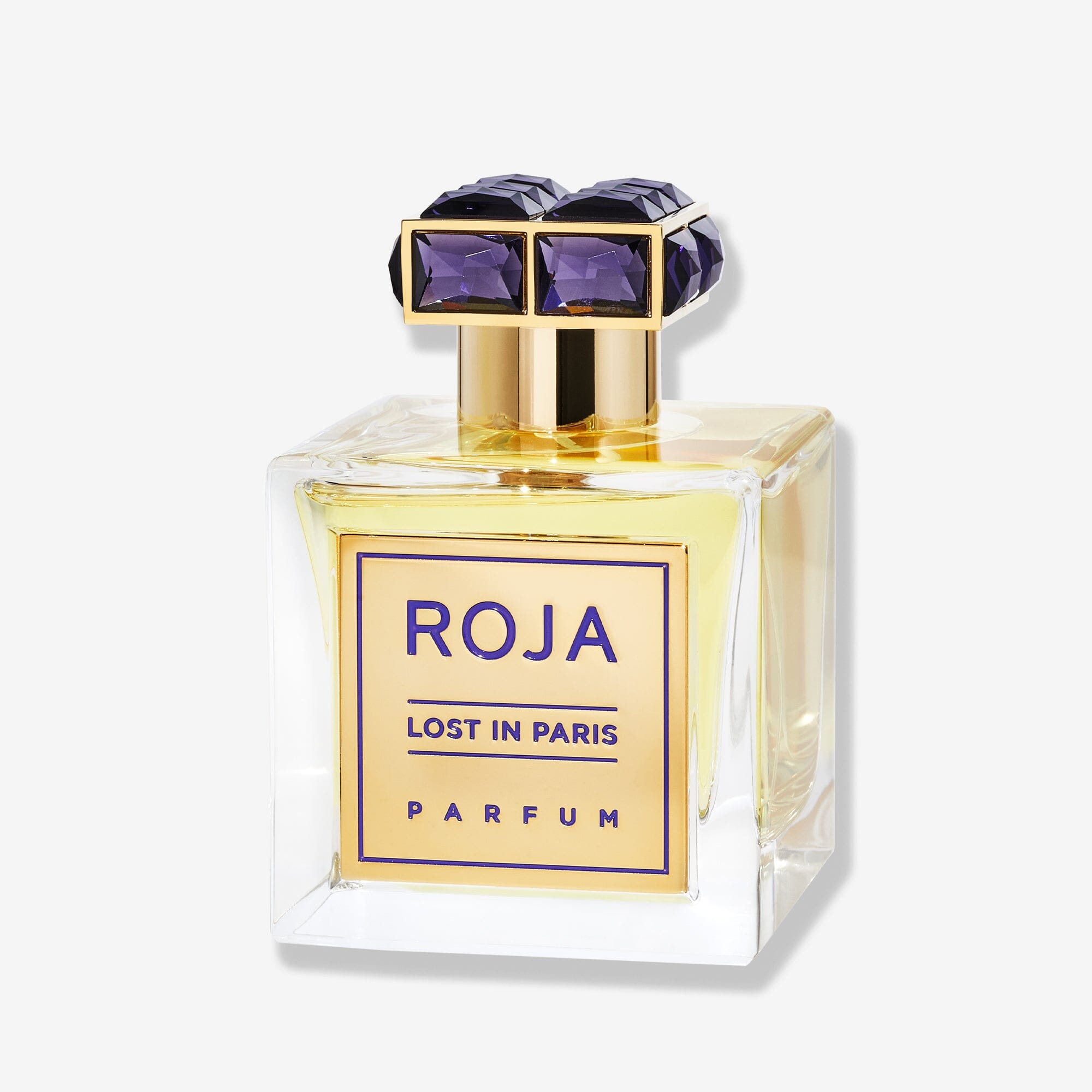 A square glass bottle of Roja Parfums Lost In Paris, a luxury fragrance with golden liquid. It has a gold label and a decorative gold cap set with large, faceted purple gems—capturing the elegance of Parisian high society.