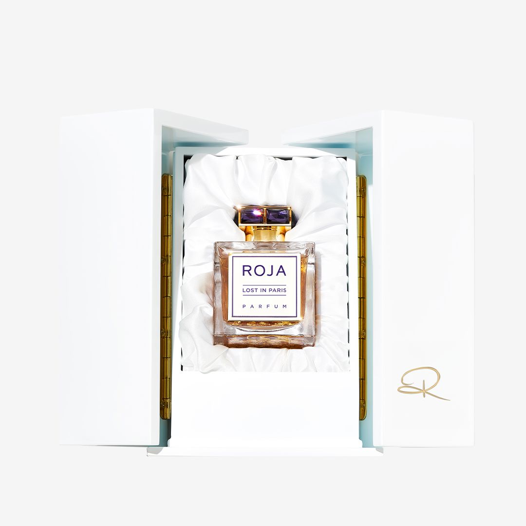 A bottle of Roja Parfums Lost In Paris is displayed upright in a white satin-lined box with gold accents and a stylized R logo on the inner panel.