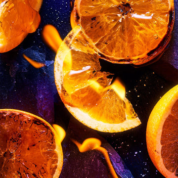 A close-up of sliced oranges with charred edges on a dark surface, bright flames evoking gourmand accords and vibrant contrast—an image capturing the dramatic character of Lost in Paris by Roja Parfums.